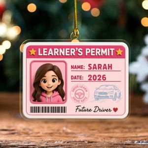 May include: A rectangular acrylic ornament shaped like a learner's permit, featuring a pink background, a cartoon portrait of a person with brown hair in a pink hoodie, the text "LEARNER'S PERMIT", "NAME: SARAH", "DATE: 2026", and "Future Driver".