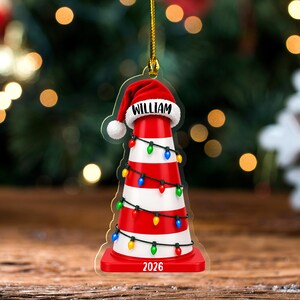 May include: A flat, acrylic Christmas tree ornament shaped like a traffic cone with red and white horizontal stripes. The cone wears a red Santa hat with the name WILLIAM printed on the brim and is wrapped in colorful string lights with 2026 at the base.