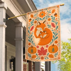May include: A rectangular garden flag featuring a central illustration of an adult fox and a kit curled together. The design includes orange pumpkins, red maple leaves, acorns, and teal floral vines on a cream background, hanging from a metal pole.