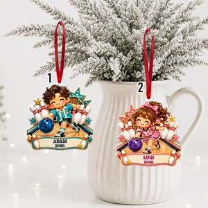 May include: Two hanging ornaments featuring cartoon characters bowling. Ornament 1 shows a character in a blue shirt with the text ADAM 2026. Ornament 2 shows a character in a pink outfit with the text LUGI 2026. Both have red ribbons.