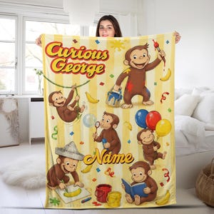 May include: A yellow and white striped fleece blanket featuring Curious George illustrations, including the character painting, reading, and holding balloons. The text reads Curious George and Name, surrounded by colorful confetti and banana graphics.
