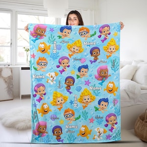 May include: A light blue fleece blanket featuring a repeating pattern of Bubble Guppies characters, including merfolk and a puppy, interspersed with bubbles, starfish, and seaweed. The word Name is printed in black text throughout the design.