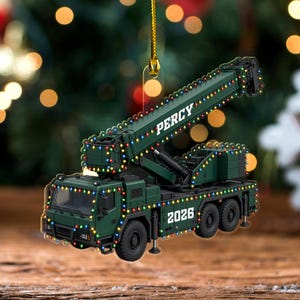 May include: A dark green mobile crane ornament hanging from a gold string. The crane is decorated with colorful Christmas lights and features the name PERCY printed in white on the boom and the year 2026 printed in white on the side panel.