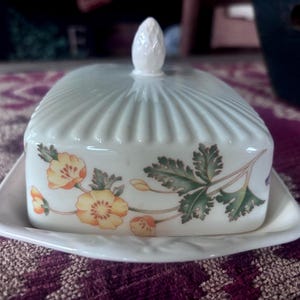 May include: A rectangular white ceramic butter dish with a ribbed lid and a pointed finial. The sides of the lid feature a botanical illustration of yellow flowers and green leaves. The base is a matching white tray with a slightly raised edge.