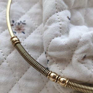 Peut inclure: Un collier en métal doré composé d'une section de fil lisse et incurvé reliée à une partie flexible en forme de ressort. Le design comprend deux petites perles sphériques polies qui séparent les éléments métalliques enroulés du bijou.