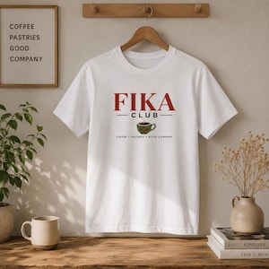 Pode incluir: Uma t-shirt branca de manga curta com o texto vermelho FIKA CLUB, um gráfico de uma chávena de café verde e o texto preto COFFEE PASTRIES GOOD COMPANY. A t-shirt está exposta num cabide de madeira contra uma parede neutra.