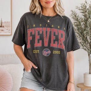 May include: A charcoal gray short-sleeve t-shirt featuring the text INDIANA FEVER in large red block letters. Below the text, the shirt displays the words EST. 2000 in yellow, flanking a circular logo with the team name and a basketball graphic.