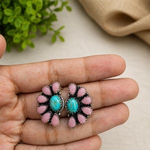 May include: A pair of stud earrings featuring a central oval turquoise stone surrounded by a fan-shaped arrangement of small, smooth, pink oval stones set in a silver-toned metal frame with a textured, oxidised finish around the edges.