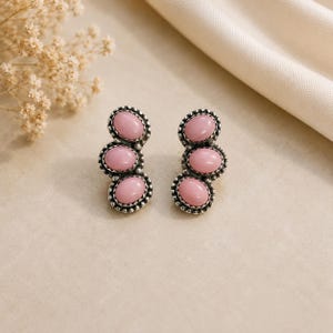 Può includere: Un paio di orecchini a lobo con tre pietre rosa di forma ovale incastonate in argento ossidato. Ogni pietra è circondata da un bordo in metallo perlato, creando un design a grappolo verticale. Gli orecchini hanno una finitura metallica.