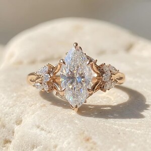 Könnte beinhalten: Ein Ring aus Roségold mit einem großen, im Marquise-Schliff facettierten Mittelstein, der von Krappen gehalten wird. Die Ringschiene ist mit kleineren Marquise- und Rundschliff-Steinen besetzt, die beidseitig eine blütenartige Anordnung bilden.