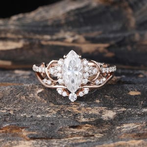 May include: A rose gold ring featuring a central marquise-cut white gemstone surrounded by a halo of smaller round white stones. The band has an intricate, swirling metal design accented with additional small round white stones along the sides.