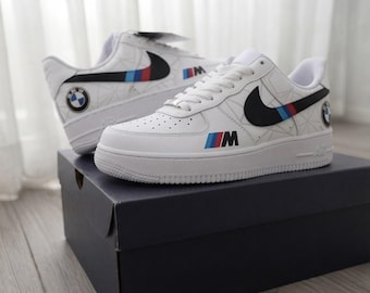 May include: A pair of white trainers featuring a black Nike swoosh with blue and red stripes, a BMW logo on the heel, and an M logo on the side. The shoes have a white geometric pattern on the upper, white laces, and are placed on a dark blue box.