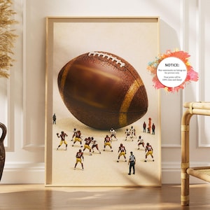 Op de afbeelding: Een verticale wandkunstprint met een grote bruine American football boven een groep miniatuur footballspelers in bordeauxrode en gouden uniformen op een zanderig veld. De print bevat een cirkelvormige kennisgeving met tekst.