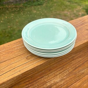 May include: A stack of four round, light mint green ceramic plates with a smooth, glossy finish. Each plate features a simple, clean design with a slightly raised rim, creating a uniform and consistent appearance for the entire set.