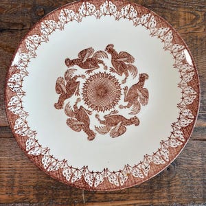 May include: A round ceramic plate with a cream-colored base and a brown transferware pattern. The center features a circular arrangement of six roosters surrounding a sunburst motif, while the rim displays a repeating leaf design.