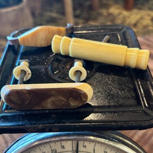 May include: Three vintage drawer pulls with cream and brown marbled resin accents. One pull is a long cylindrical shape, while the other two feature flat, rectangular designs with metal mounting screws and small circular washers attached.