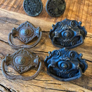 Könnte beinhalten: Eine Sammlung von sechs Möbelbeschlägen aus dunklem Metall, bestehend aus zwei runden Knöpfen mit floralen Reliefmustern und vier verzierten Schubladengriffen mit schwingenden Bügeln, die mit filigranen Schnörkeln und zentralen Medaillons versehen sind.