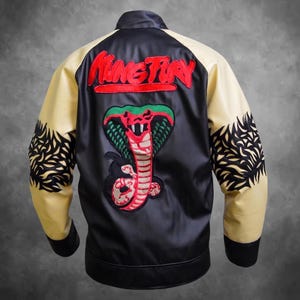 Puede incluir: Una chaqueta de satén negra y color crema con una cobra grande bordada en la espalda en tonos verdes, rojos y negros. Sobre la cobra, el texto "KUNG FURY" está bordado en rojo. Las mangas tienen patrones negros en forma de llamas.