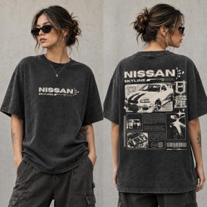 Op de afbeelding: Een antracietgrijs T-shirt met korte mouwen en een vintage-gewassen afwerking. De voorkant toont de tekst NISSAN SKYLINE. De achterkant bevat een grote afbeelding van een Nissan Skyline R34 auto, technische tekst en Japanse tekens in wit.