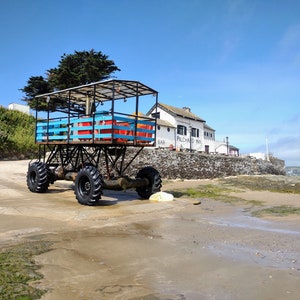 May include: A blue and orange metal cart with large wheels sits on a sandy beach in front of a white building with the words "Pilchard Inn" on the side.
