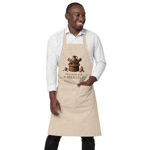 May include: A beige apron featuring a graphic of hands shaping a clay pot on a pottery wheel, with smaller pottery items nearby. Below the graphic, the text reads "These hands work MIRACLES" in brown lettering, accompanied by small leaf illustrations.