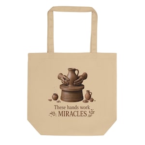 Peut inclure: Un sac fourre-tout beige avec un motif marron représentant deux mains façonnant une cruche en argile sur un tour de potier. De petits objets en poterie entourent la base. Sous l'illustration, le texte indique : These hands work MIRACLES.