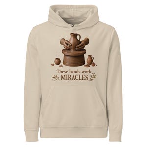 May include: A beige hooded sweatshirt featuring a graphic of two hands shaping a clay pitcher on a pottery wheel. Smaller clay vessels surround the wheel. Below the graphic, the text reads: These hands work MIRACLES, flanked by small leaf sprigs.