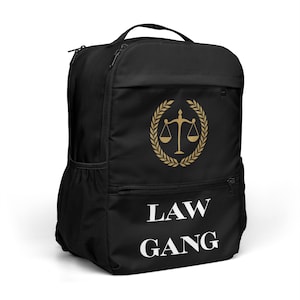 May include: A black backpack featuring a gold-colored graphic of a scale of justice inside a laurel wreath. Below the graphic, the text "LAW GANG" is printed in white capital letters. The backpack includes a top handle and a front zippered pocket.