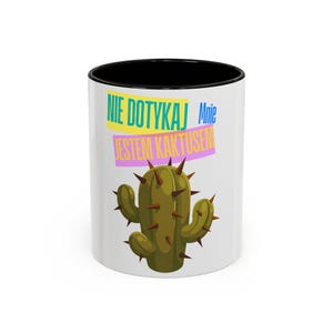 May include: A white ceramic mug with a black interior and rim. The exterior features a green cactus illustration with brown spikes and the Polish text "NIE DOTYKAJ Mnie JESTEM KAKTUSEM" printed in blue, yellow, and pink rectangular blocks.