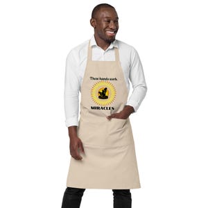 May include: A beige apron featuring a central graphic of a yellow sunburst with a black silhouette of hands working on a pottery wheel. The text above the graphic reads "These hands work" and the text below the graphic reads "MIRACLES".