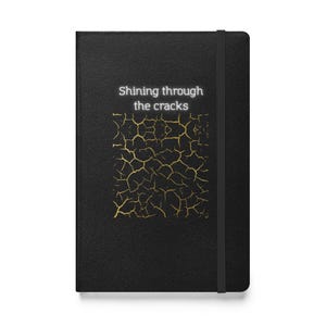 May include: A black hardcover notebook with an elastic closure band. The front cover features the white text "Shining through the cracks" above a graphic pattern of gold-colored lines resembling a cracked surface texture.