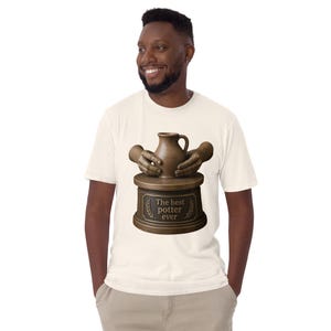 May include: A cream-colored t-shirt featuring a graphic of two bronze-toned hands shaping a clay pitcher on a pottery wheel. The base of the wheel includes a plaque with the text "The best potter ever" flanked by two laurel leaf icons.