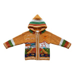 Op de afbeelding: Een mosterdgeel gebreid vest met capuchon en een ritssluiting over de gehele lengte. Het kledingstuk toont een kleurrijk landschap met een zon, vogels, een berg en kleine dierfiguren. De mouwen hebben horizontale groene en witte strepen.