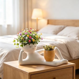 May include: A white ceramic planter shaped like a reclining figure with closed eyes and a smile. The head holds a cluster of small, colorful flowers, while a separate, smaller yellow pot containing a green succulent rests on the figure's legs.