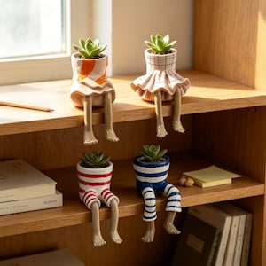 May include: Four ceramic planters shaped like figures with dangling legs, each holding a small green succulent. Two planters feature skirt-like bases with orange or blue grid patterns, while two feature pant-like bases with red or blue horizontal stripes.
