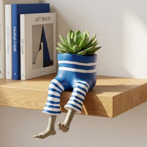Op de afbeelding: Een blauw-witte gestreepte plantenpot in de vorm van een zittend figuur met bungelende benen, met daarin een groene vetplant. De pot heeft horizontale strepen op de romp en benen, met voeten in een neutrale kleur die naar beneden wijzen.