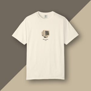 May include: A cream-colored short-sleeve t-shirt featuring a small graphic of a vintage beige computer monitor centered on the chest. Below the monitor, the text reads All-In-One. 1986 in a simple black font. The shirt has a crew neckline.