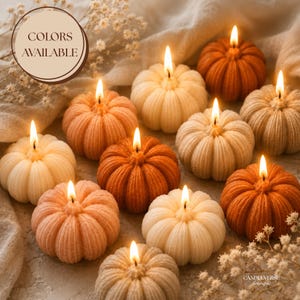 May include: A collection of ten lit pumpkin-shaped candles in various shades of orange, cream, and beige. Each candle features a ribbed texture and a single burning wick. A circular graphic with the text COLORS AVAILABLE is in the upper left.