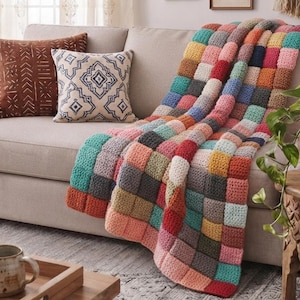 May include: A handmade crochet throw blanket featuring a multi-colored patchwork design composed of small, square-shaped yarn blocks in various shades including teal, mustard yellow, coral, grey, pink, and blue, draped over a neutral-toned sofa.