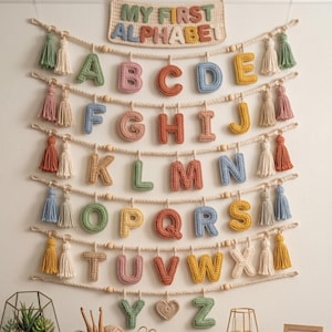 May include: A wall hanging featuring the text MY FIRST ALPHABET in crocheted letters above five rows of rope strung with wooden beads and colorful crocheted letters from A to Z. Each row is accented with decorative yarn tassels in various colors.