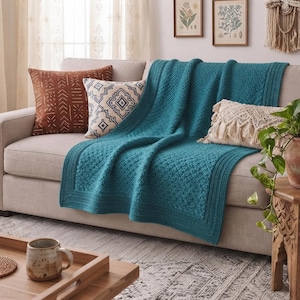 May include: A teal knitted throw blanket with a textured diamond pattern and a ribbed border, draped over a light beige sofa. The blanket is accompanied by a rust-colored patterned pillow, a white pillow with blue geometric designs, and a cream fringe pillow.