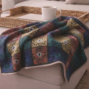 May include: A woven throw blanket featuring a repeating grid pattern of squares in deep shades of blue, green, yellow, and orange. Each square contains a stylised eye motif with intricate geometric detailing and a light beige finished edge.