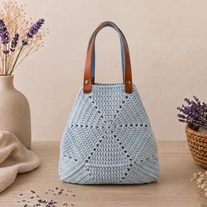 May include: A light blue crochet handbag featuring a geometric star-like pattern throughout the body. The bag has two brown leather handles attached with silver-toned metal rivets at the top, providing a structured shape for the soft textile.