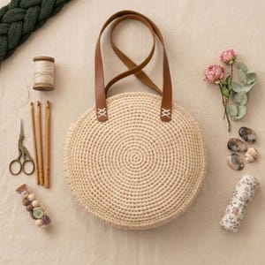 May include: A round beige crochet bag with brown leather straps, accompanied by a wooden spool of thread, three wooden crochet hooks, metal scissors, a small container of buttons, pink flowers, smooth stones, and a roll of floral fabric.
