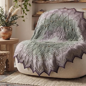 May include: A round crochet blanket featuring a complex mandala pattern in gradient shades of sage green and dusty purple. The blanket has a textured surface with intricate stitch work and a pointed, scalloped edge that drapes over a cream-colored chair.