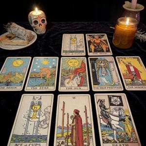 May include: A collection of tarot cards arranged on a dark surface, including The Moon, The Star, The Sun, The High Priestess, The Magician, The Devil, and Ace of Cups. A lit candle in a skull-shaped holder and a jar candle are also visible.