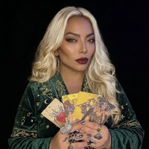 May include: A dark green velvet robe featuring gold embroidered celestial motifs including stars and moons. The garment is worn with multiple silver-toned rings on the fingers, while four tarot cards are held in the hands, displaying various illustrations.