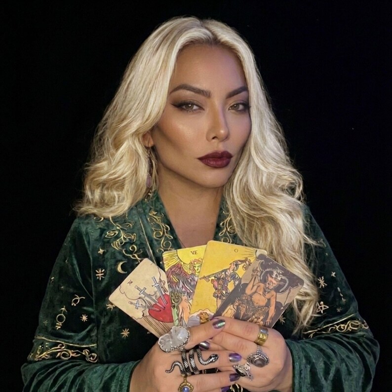 May include: A dark green velvet robe featuring gold embroidered celestial motifs including stars and moons. The garment is worn with multiple silver-toned rings on the fingers, while four tarot cards are held in the hands, displaying various illustrations.