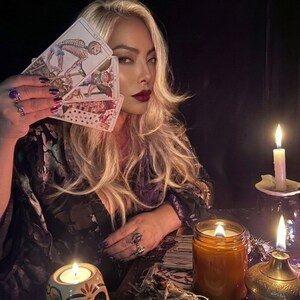 May include: A set of three tarot cards held in a hand, featuring illustrations of a skeleton, a figure with wings, and a collection of coins. The cards are displayed alongside several lit candles in glass jars and a metal oil lamp on a dark surface.