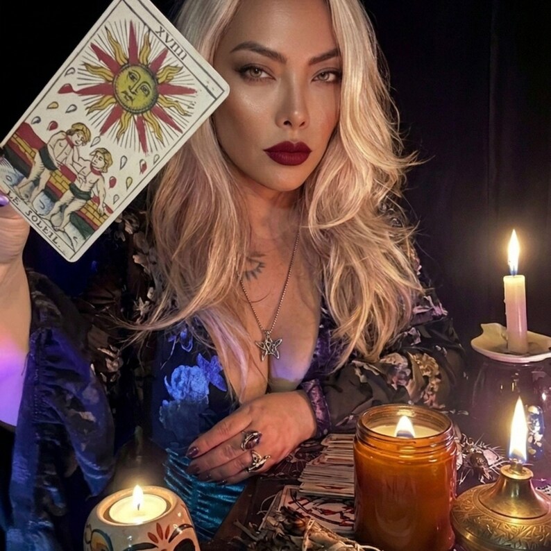 May include: A rectangular tarot card titled Le Soleil XVIII featuring a yellow sun with a face, red and orange rays, and two figures standing before a brick wall. The card is held up against a dark background with lit candles and a star pendant.